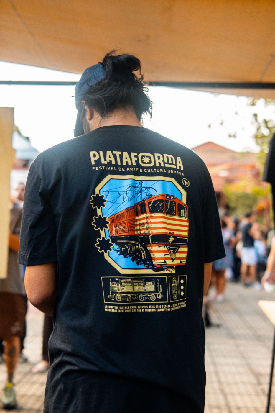 Camiseta preta do festival Plataforma, com estampa de locomotiva em várias cores, vista de costas